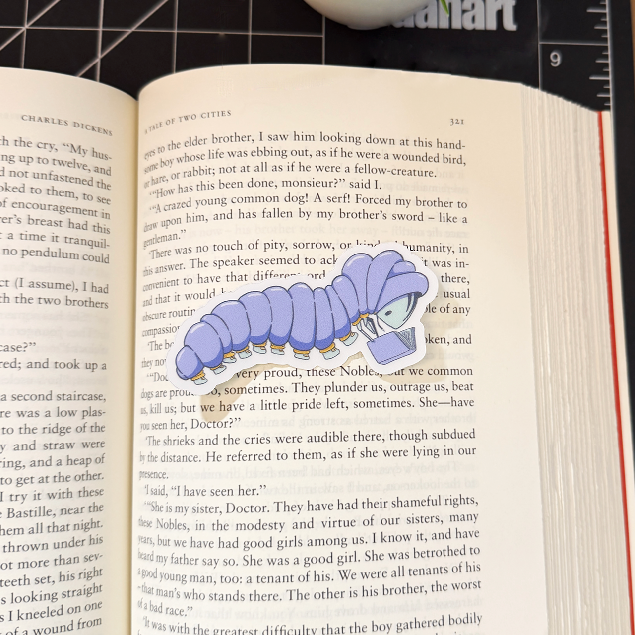 Sticker of a caterpillar wearing a purple puffy jacket with yellow cuffs and light blue shoes. They are reading a purple book. This sticker sits on a book with text so that the scale of the sticker can be seen.