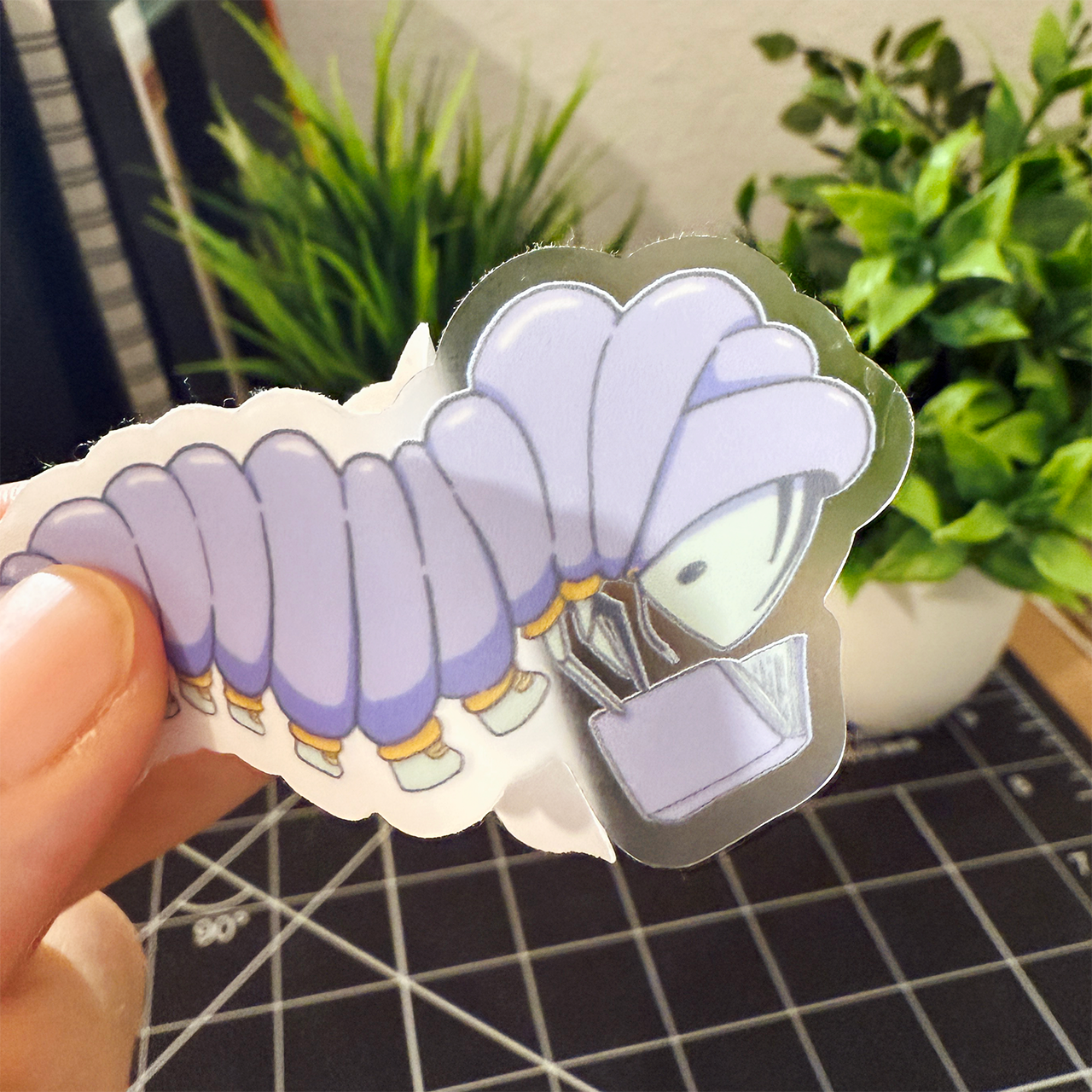 Hand holding a sticker of a caterpillar wearing a purple puffy jacket with yellow cuffs and light blue shoes. They are reading a purple book. The sticker backing is peeled off halfway so you can see the sticker has a clear background and it is being held in front of a desk with plants.