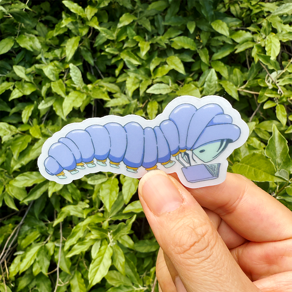 Hand holding a sticker of a caterpillar wearing a purple puffy jacket with yellow cuffs and light blue shoes. They are reading a purple book. This sticker is held in front of a background of green leaves.