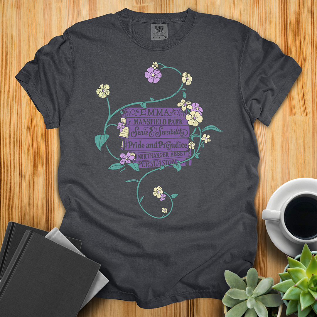 Graphite Gray Comfort Colors T-shirt featuring all of Jane Austen's novels surrounded by a delicate floral design with white and purple flowers and green vines. The shirt sits on a wood floor with plants, coffee, and books placed around it.