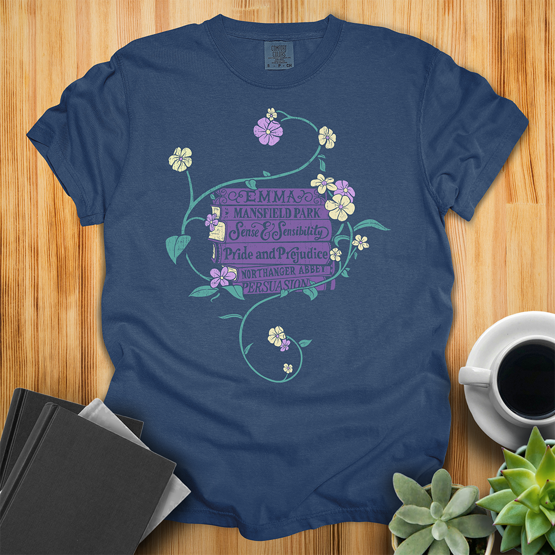 Navy Blue Comfort Colors T-shirt featuring all of Jane Austen's novels surrounded by a delicate floral design with white and purple flowers and green vines. The shirt sits on a wood floor with plants, coffee, and books placed around it.