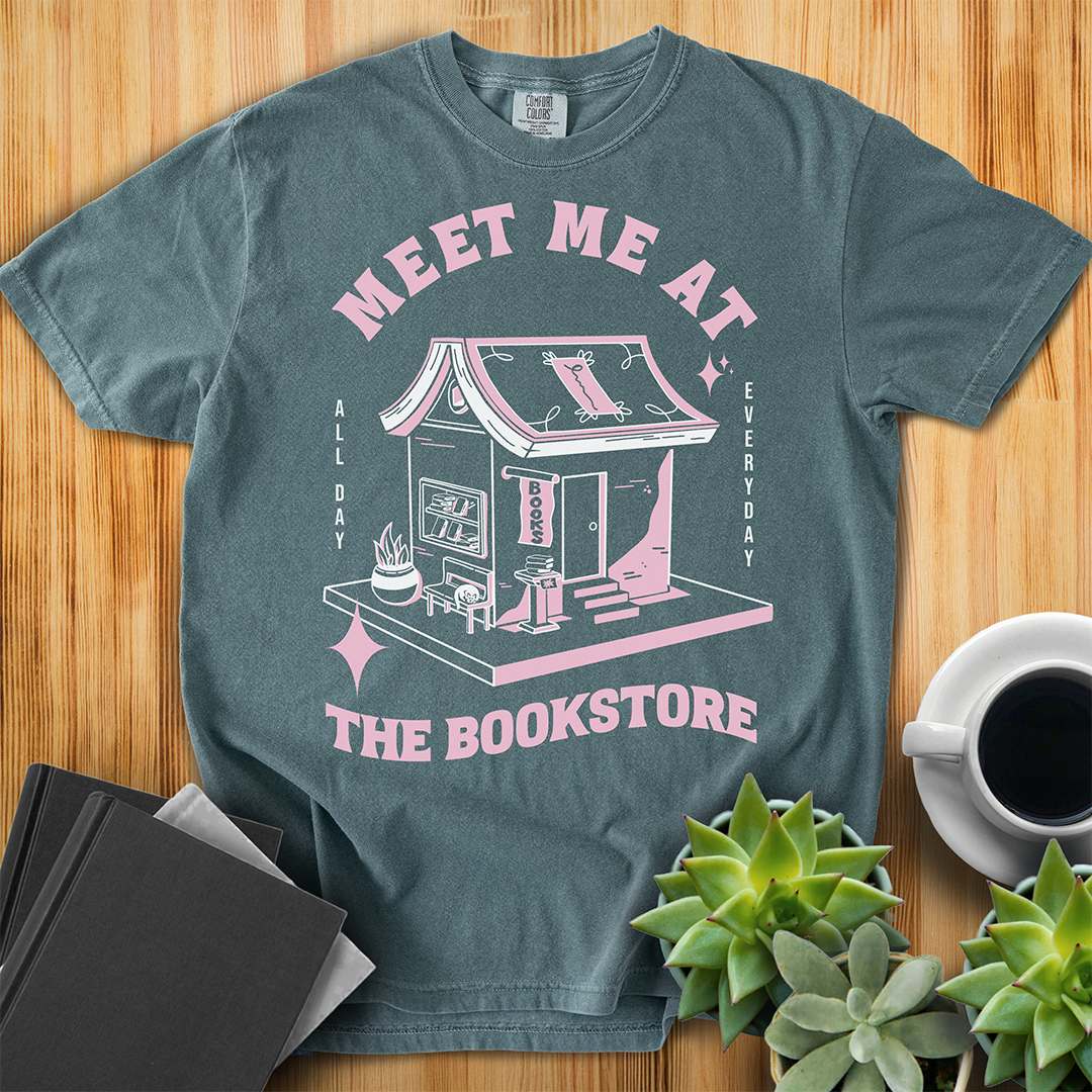 Blue Spruce Comfort Colors t-shirt with a hand-drawn design of a bookstore with a cute cat on a bench and books in the window, and an open book as a roof, with the text "Meet me at the Bookstore", Vertical text on either side reads all day, and everyday. The shirt sits on a wood floor with plants, coffee, and books placed around it.