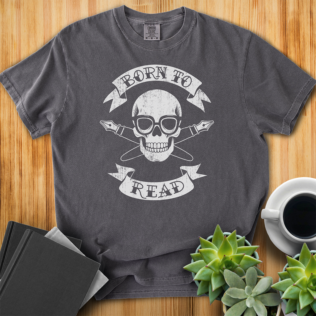 Pepper Gray Comfort Colors T-shirt with a hand-drawn illustration of a skull and crossbones, except instead of crossbones, it has crossed pens and he's wearing nerdy glasses. The text is in a banner on top and bottom that reads "Born to Read" The shirt sits on a wood floor with plants, coffee, and books placed around it.