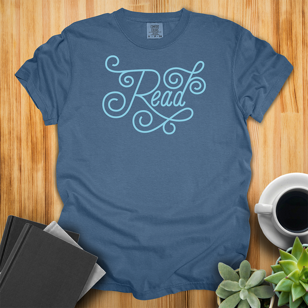 Midnight Blue Comfort Colors T-shirt featuring the word "read" in hand-lettered light blue script. The shirt sits on a wood floor with plants, coffee, and books placed around it.