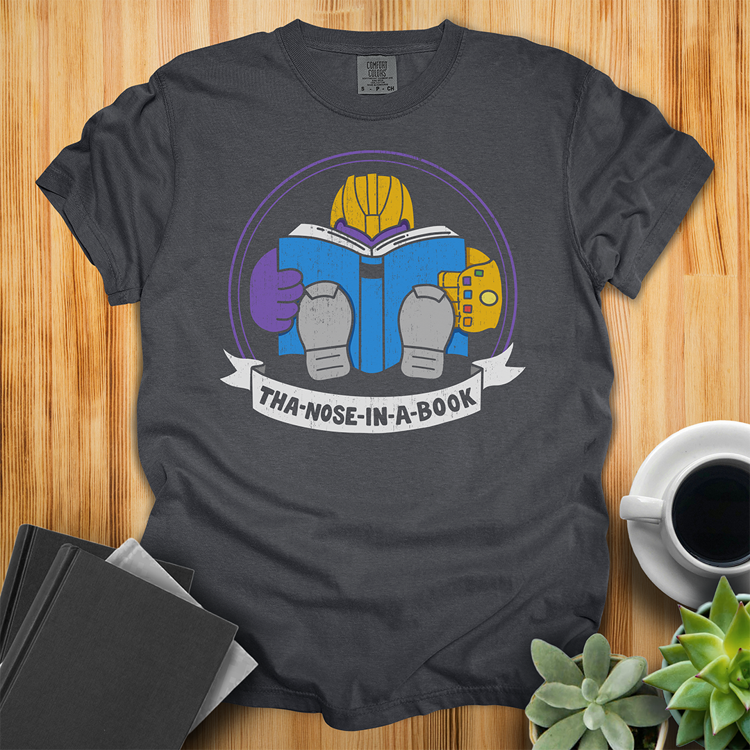 Graphite Comfort Colors t-shirt with a purple villain wearing a gold glove with gemstones. He is reading a blue book and the bottom banner reads "Tha-nose-in-a-book" as a parody. The shirt sits on a wood floor with plants, coffee, and books placed around it.