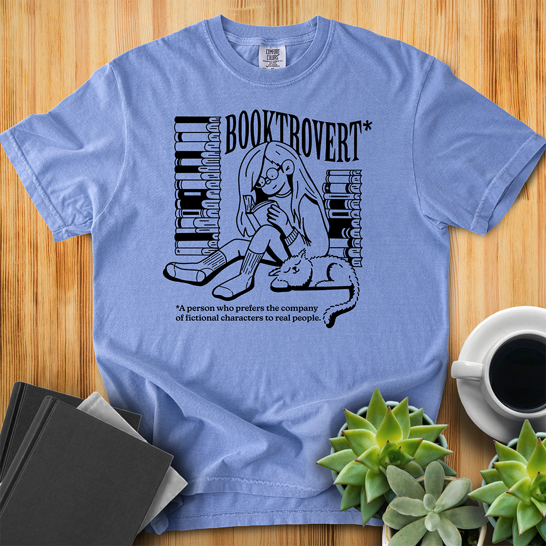 Washed Denim Blue Comfort Colors t-shirt with the text "Booktrovert." A woman holds a book reading, with her cat sitting next to her sleeping. Behind her are two piles of books. The bottom reads "A person who prefers the company of fictional characters to read people." The shirt sits on a wood floor with plants, coffee, and books placed around it.