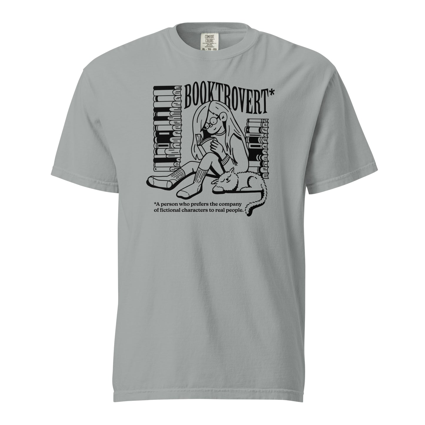 Granite Gray Comfort Colors t-shirt with the text "Booktrovert." A woman holds a book reading, with her cat sitting next to her sleeping. Behind her are two piles of books. The bottom reads "A person who prefers the company of fictional characters to read people."