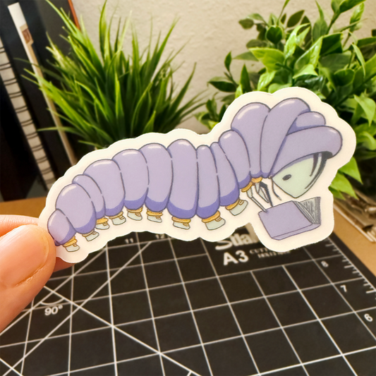 Hand holding a sticker of a caterpillar wearing a purple puffy jacket with yellow cuffs and light blue shoes. They are reading a purple book. The sticker is being held in front of a desk with green plants.