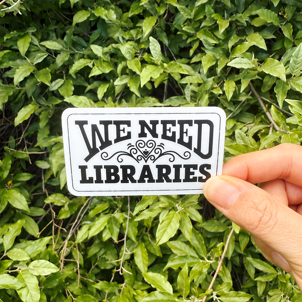 Hand holding a 'We Need Libraries' rectangular sticker with a heart flourish in front of green leaves