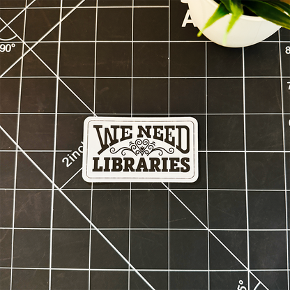 A rectangular We need libraries sticker sitting with a heart flourish in the middle, on a one inch black gridded slate so that the scale is apparent.