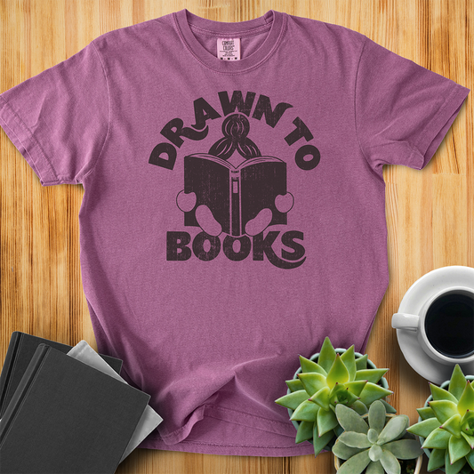 Berry Purple Pink Red t-shirt with Drawn to Books logo, text says "Drawn to Books" which is framed around a girl reading a book, and you can see only the top of her ponytail, hands and feet, and the big book she's reading. The shirt sits on a wood floor with plants, coffee, and books placed around it. 