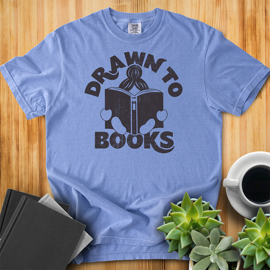 Washed Denim Blue t-shirt with Drawn to Books logo, text says "Drawn to Books" which is framed around a girl reading a book, and you can see only the top of her ponytail, hands and feet, and the big book she's reading. The shirt sits on a wood floor with plants, coffee, and books placed around it. 
