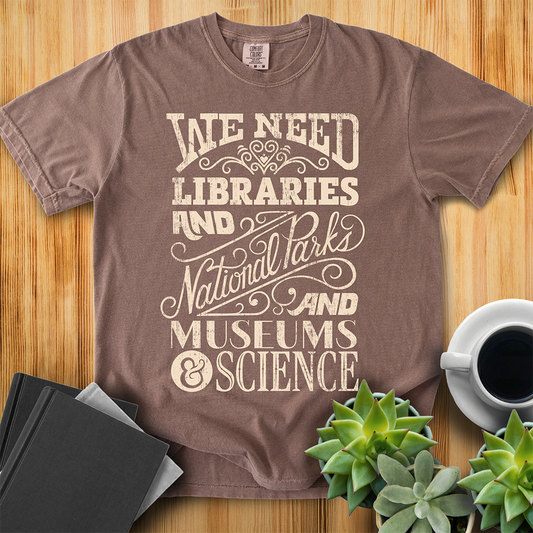 Espresso Brown Comfort Colors t-shirt with a hand lettered typography design reading "We Need Libraries and National Parks and Museums and Science." The shirt sits on a wood floor with plants, coffee, and books placed around it. 