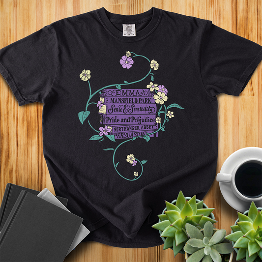 Black Comfort Colors T-shirt featuring all of Jane Austen's novels surrounded by a delicate floral design with white and purple flowers and green vines. The shirt sits on a wood floor with plants, coffee, and books placed around it. 
