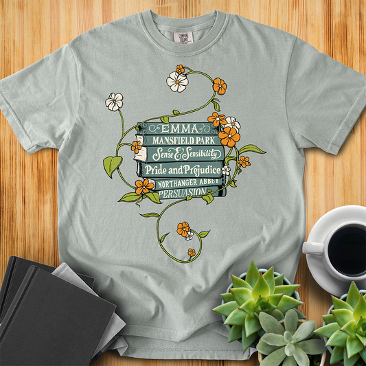 Bay Gray Comfort Colors T-shirt featuring all of Jane Austen's novels with teal covers and hand-type of each title, surrounded by a delicate floral design with white and orange flowers and green vines. The shirt sits on a wood floor with plants, coffee, and books placed around it. 