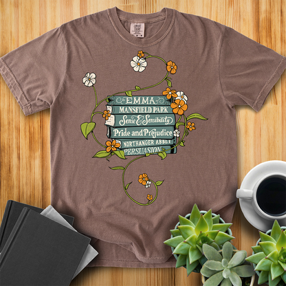 Espresso Brown Comfort Colors T-shirt featuring all of Jane Austen's novels with teal covers and hand-type of each title, surrounded by a delicate floral design with white and orange flowers and green vines. The shirt sits on a wood floor with plants, coffee, and books placed around it. 