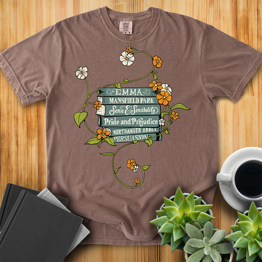 Espresso Brown Comfort Colors T-shirt featuring all of Jane Austen's novels with teal covers and hand-type of each title, surrounded by a delicate floral design with white and orange flowers and green vines. The shirt sits on a wood floor with plants, coffee, and books placed around it. 