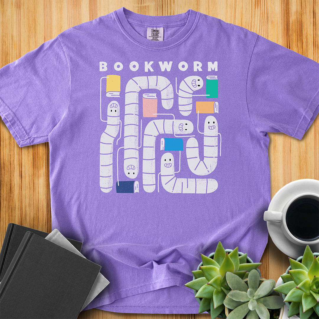 Violet purple t-shirt with 'Bookworm' design, features a hand-drawn design with white worms and happy faces . Each worm is holding up a book. The shirt sits on a wood floor with plants, coffee, and books placed around it. 