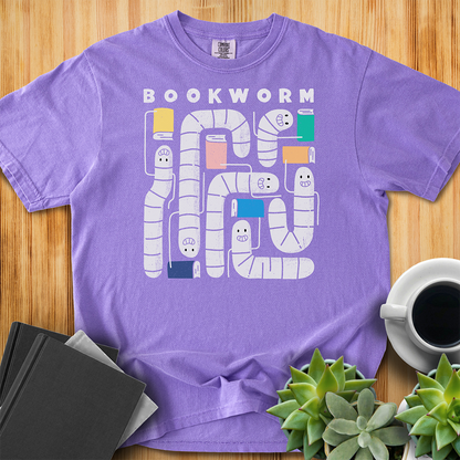 Violet purple t-shirt with 'Bookworm' design, features a hand-drawn design with white worms and happy faces . Each worm is holding up a book. The shirt sits on a wood floor with plants, coffee, and books placed around it. 
