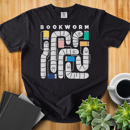 Black t-shirt with 'Bookworm' design, features a hand-drawn design with white worms and happy faces . Each worm is holding up a book. The shirt sits on a wood floor with plants, coffee, and books placed around it. 