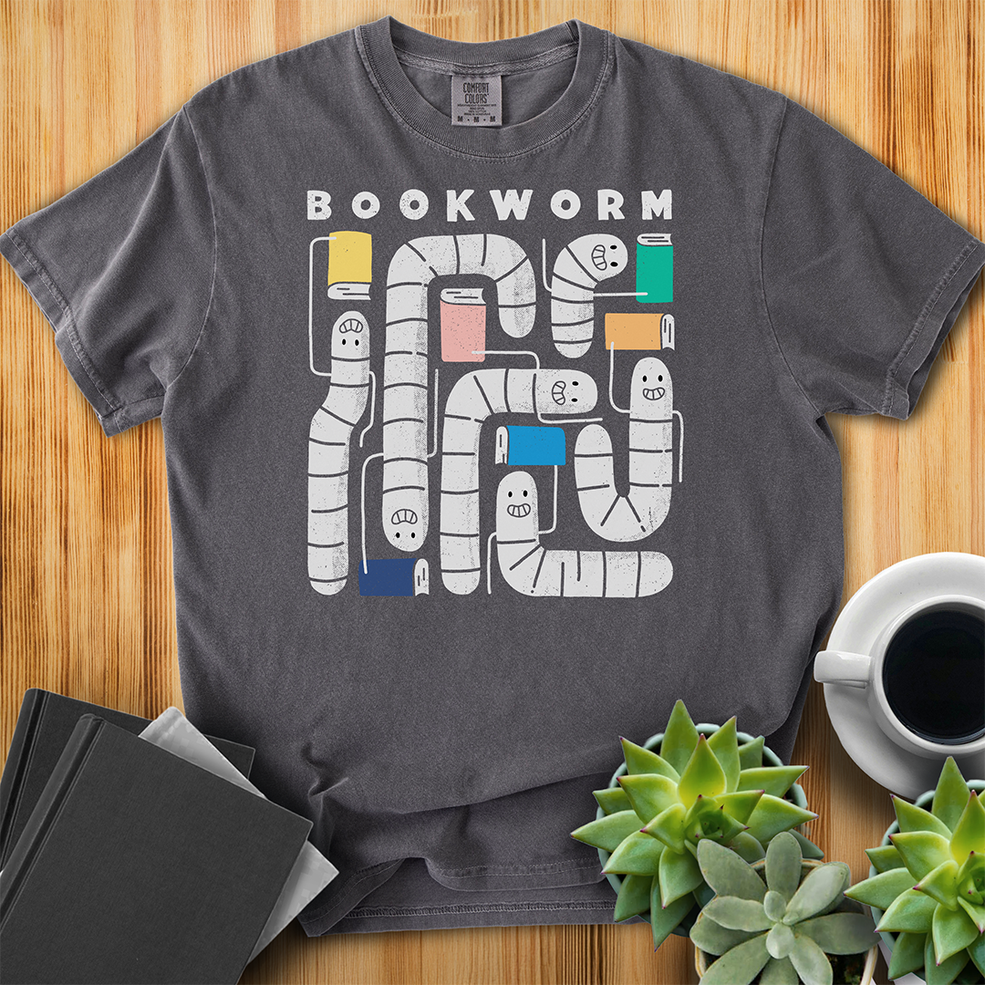 Pepper gray t-shirt with 'Bookworm' design, features a hand-drawn design with white worms and happy faces . Each worm is holding up a book. The shirt sits on a wood floor with plants, coffee, and books placed around it. 