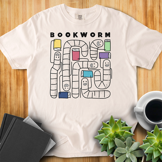 Ivory white Comfort Colors t-shirt with 'Bookworm' design, features a hand-drawn design with black outlined worms and happy faces . Each worm is holding up a book. The shirt sits on a wood floor with plants, coffee, and books placed around it. 