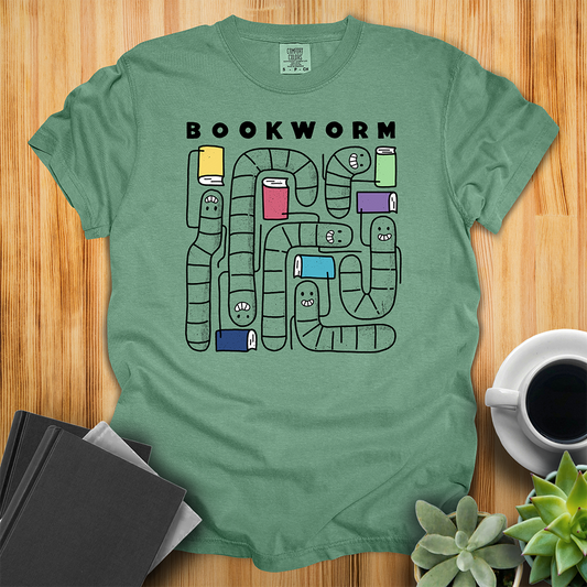Light Green Comfort Colors t-shirt with 'Bookworm' design, features a hand-drawn design with black outlined worms and happy faces . Each worm is holding up a book. The shirt sits on a wood floor with plants, coffee, and books placed around it. 