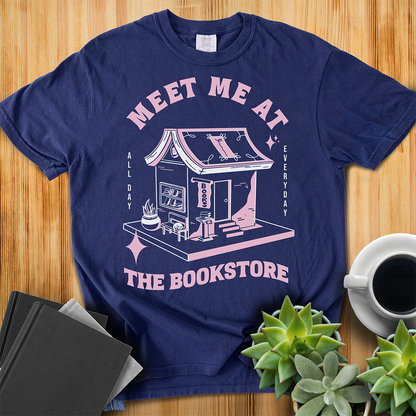 True Navy Comfort Colors t-shirt with a hand-drawn design of a bookstore with a cute cat on a bench and books in the window, and an open book as a roof, with the text "Meet me at the Bookstore", Vertical text on either side reads all day, and everyday. The shirt sits on a wood floor with plants, coffee, and books placed around it. 