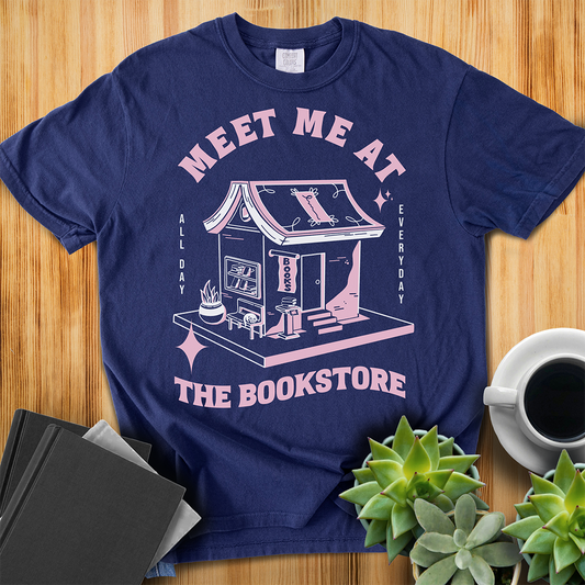 True Navy Comfort Colors t-shirt with a hand-drawn design of a bookstore with a cute cat on a bench and books in the window, and an open book as a roof, with the text "Meet me at the Bookstore", Vertical text on either side reads all day, and everyday. The shirt sits on a wood floor with plants, coffee, and books placed around it. 