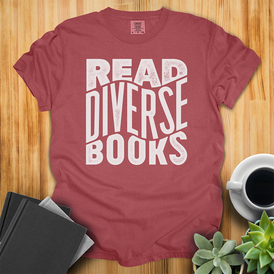 
Brick Red t-shirt with hand lettered design that says "Read Diverse Books" in white text. The shirt sits on a wood floor with plants, coffee, and books placed around it.