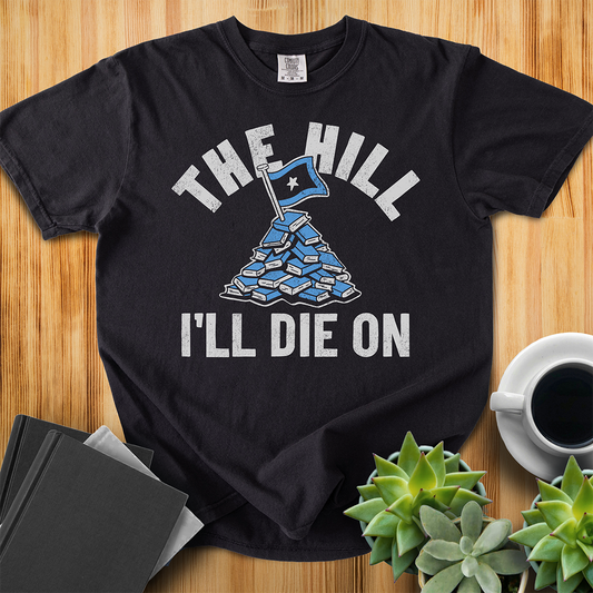 Black Comfort Colors T-shirt with a hand-drawn illustration of a light blue stack of books with the phrase "The Hill I'll Die On." The shirt sits on a wood floor with plants, coffee, and books placed around it. 