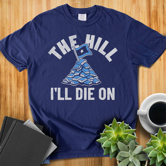 True Navy Blue Comfort Colors T-shirt with a hand-drawn illustration of a light blue stack of books with the phrase "The Hill I'll Die On." The shirt sits on a wood floor with plants, coffee, and books placed around it. 