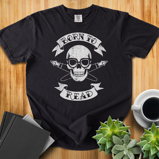 Black Comfort Colors T-shirt with a hand-drawn illustration of a skull and crossbones, except instead of crossbones, it has crossed pens and he's wearing nerdy glasses. The text is in a banner on top and bottom that reads "Born to Read" The shirt sits on a wood floor with plants, coffee, and books placed around it. 
  