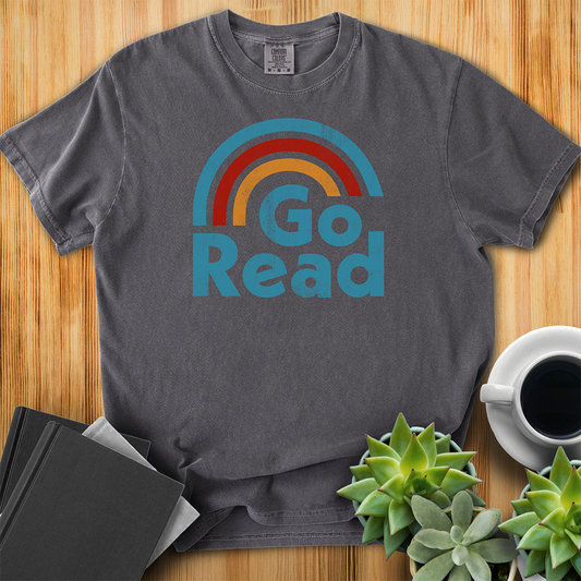 Pepper Gray Comfort Colors T-shirt with the phrase "go read" and a three-color rainbow of blue red and yellow. It has grit and a retro feel. The shirt sits on a wood floor with plants, coffee, and books placed around it. 