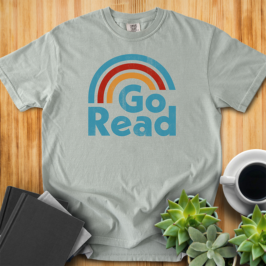 Bay Gray Comfort Colors T-shirt with the phrase "go read" and a three-color rainbow of blue red and yellow. It has grit and a retro feel. The shirt sits on a wood floor with plants, coffee, and books placed around it. 