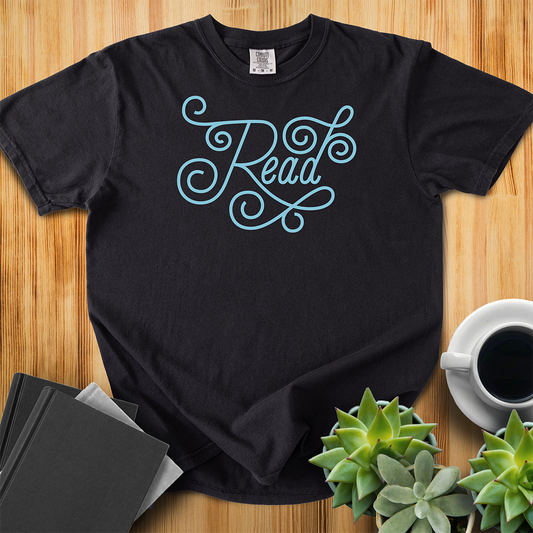 Black Comfort Colors T-shirt featuring the word "read" in hand-lettered light blue script. The shirt sits on a wood floor with plants, coffee, and books placed around it. 
