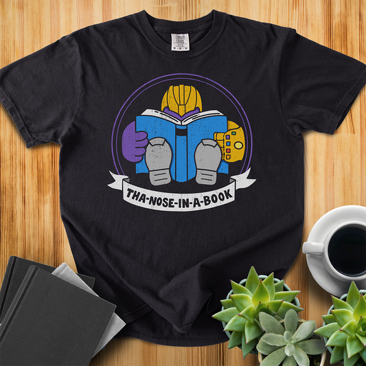 Black Comfort Colors t-shirt with a purple villain wearing a gold glove with gemstones. He is reading a blue book and the bottom banner reads "Tha-nose-in-a-book" as a parody. The shirt sits on a wood floor with plants, coffee, and books placed around it. 