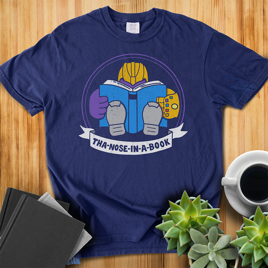True Navy Blue Comfort Colors t-shirt with a purple villain wearing a gold glove with gemstones. He is reading a blue book and the bottom banner reads "Tha-nose-in-a-book" as a parody. The shirt sits on a wood floor with plants, coffee, and books placed around it. 