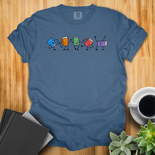 Midnight Blue Comfort Colors T-shirt with colorful dancing hand-drawn book characters. Each one is in a different dance pose with different happy facial expressions. The shirt sits on a wood floor with plants, coffee, and books placed around it. 