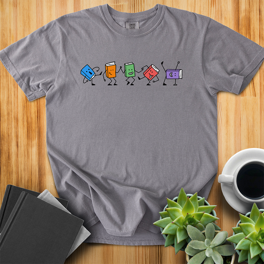Grey Comfort Colors T-shirt with colorful dancing hand-drawn book characters. Each one is in a different dance pose with different happy facial expressions. The shirt sits on a wood floor with plants, coffee, and books placed around it. 