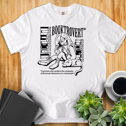 White Comfort Colors t-shirt with the text "Booktrovert." A woman holds a book reading, with her cat sitting next to her sleeping. Behind her are two piles of books. The bottom reads "A person who prefers the company of fictional characters to read people." The shirt sits on a wood floor with plants, coffee, and books placed around it. 