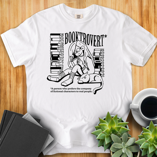 White Comfort Colors t-shirt with the text "Booktrovert." A woman holds a book reading, with her cat sitting next to her sleeping. Behind her are two piles of books. The bottom reads "A person who prefers the company of fictional characters to read people." The shirt sits on a wood floor with plants, coffee, and books placed around it. 