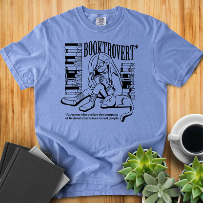 Washed Denim Blue Comfort Colors t-shirt with the text "Booktrovert." A woman holds a book reading, with her cat sitting next to her sleeping. Behind her are two piles of books. The bottom reads "A person who prefers the company of fictional characters to read people." The shirt sits on a wood floor with plants, coffee, and books placed around it. 
