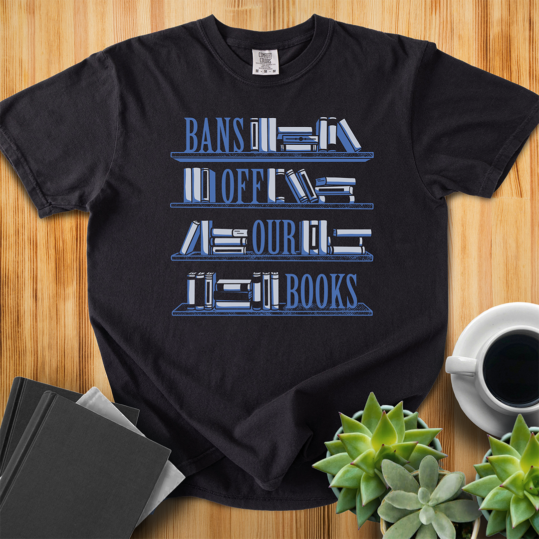 Black Comfort Colors t-shirt with Book Bans design, features a hand-drawn bookshelf design with the words "Bans off our Books," one word sits on each of the shelves. The shirt sits on a wood floor with plants, coffee, and books placed around it. 
