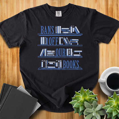 Black Comfort Colors t-shirt with Book Bans design, features a hand-drawn bookshelf design with the words "Bans off our Books," one word sits on each of the shelves. The shirt sits on a wood floor with plants, coffee, and books placed around it. 