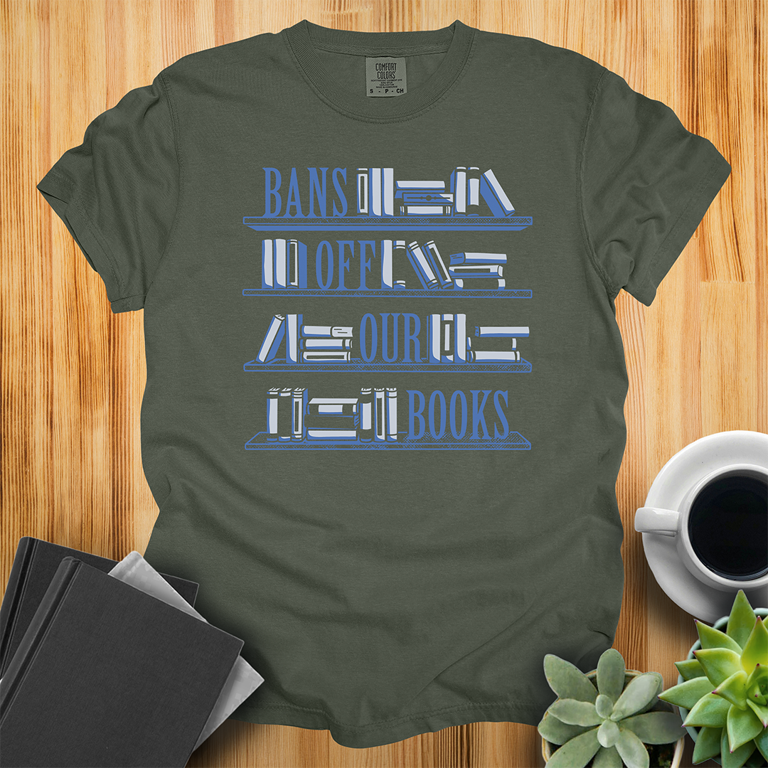 Sage Green Comfort Colors t-shirt with Book Bans design, features a hand-drawn bookshelf design with the words "Bans off our Books," one word sits on each of the shelves. The shirt sits on a wood floor with plants, coffee, and books placed around it. 