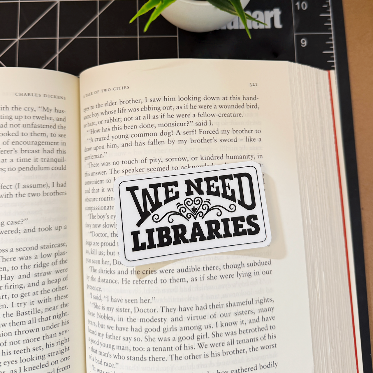 A white 'We Need Libraries' rectangular sticker with a heart flourish sitting on a book for scale.