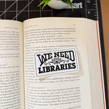 A white 'We Need Libraries' rectangular sticker with a heart flourish sitting on a book for scale.