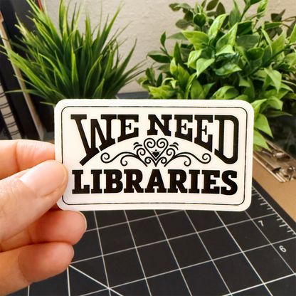 Hand holding a 'We Need Libraries' rectangular sticker with a heart flourish, plants in the background