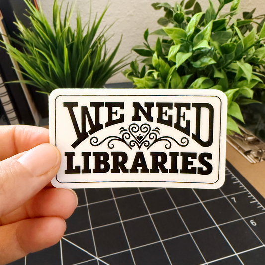 Hand holding a 'We Need Libraries' rectangular sticker with a heart flourish, plants in the background